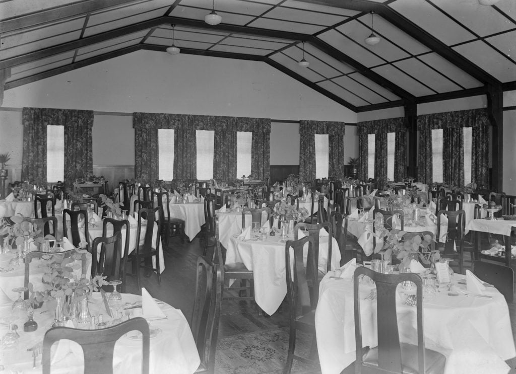 The  dining room, with round tables and high wooden chairs. The tables are covered in linen table cloths with  drinking glasses and serviettes. Large windows with curtains in the background. 