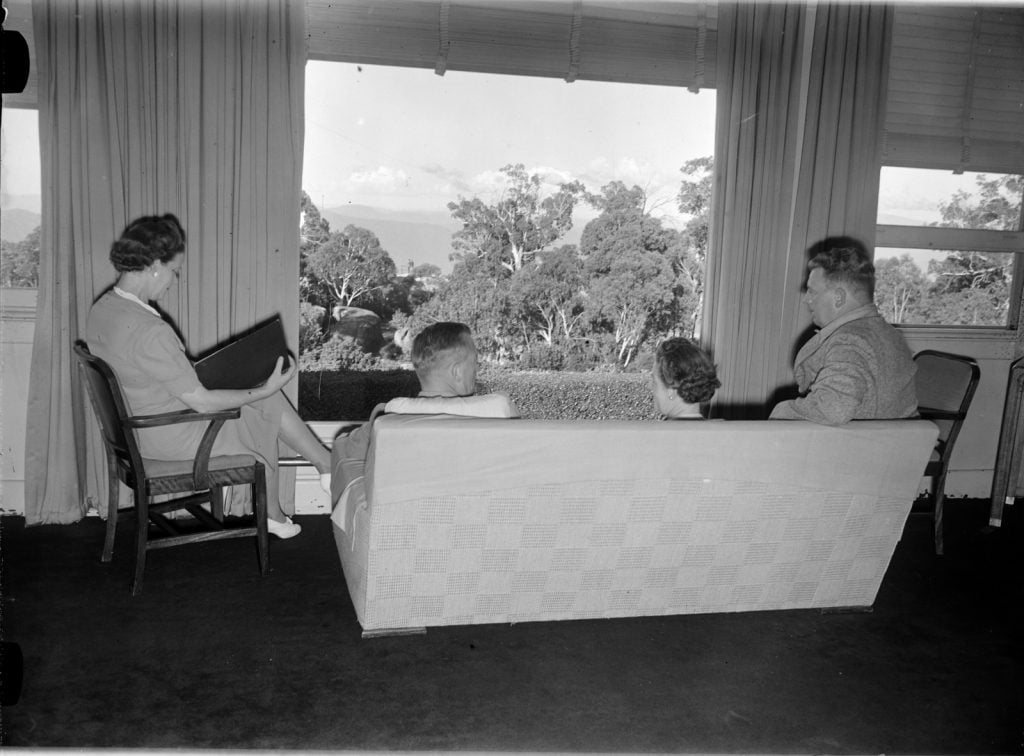 Two men and two women  sit on chairs and a sofa in front of a large window in the drawing room. The view shows large gum trees and mountains in the distance. One woman reads. 