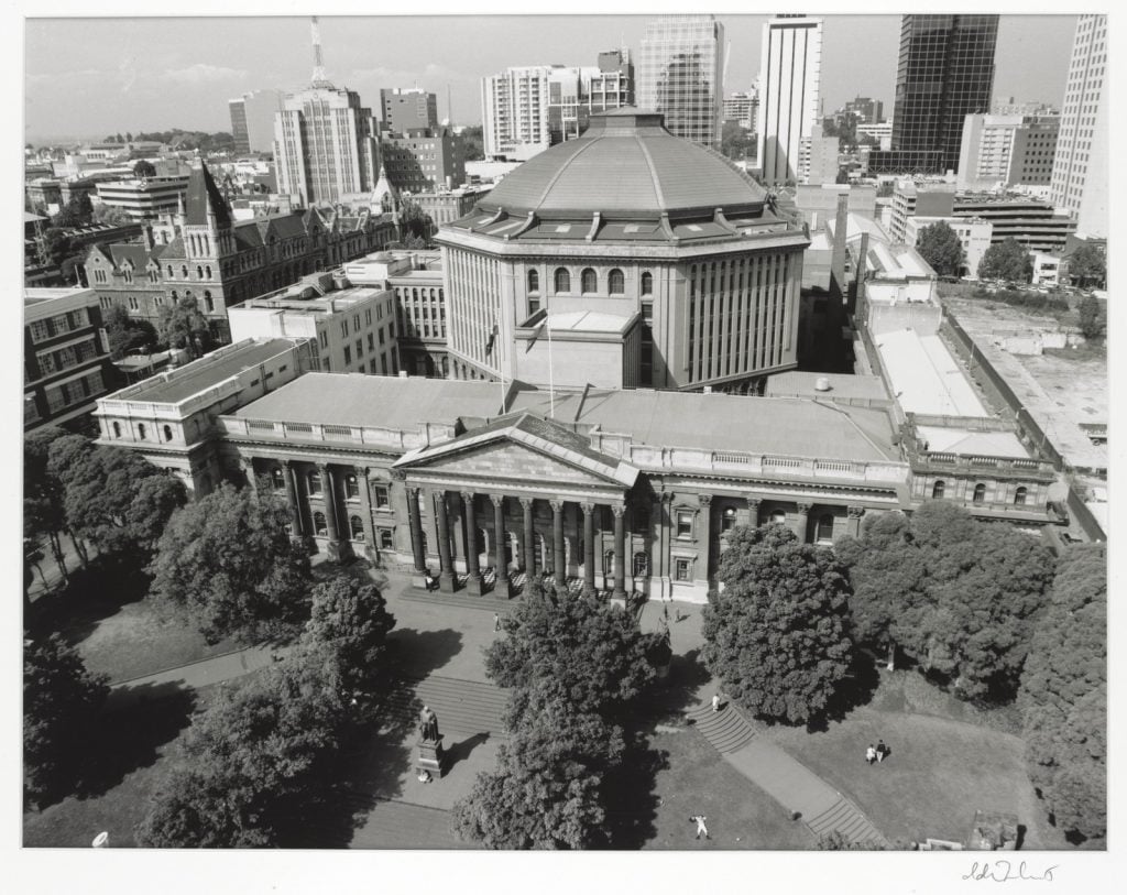 Black and white photograph of elevated view of the State Library of Victoria and surroundings, showing thick vegetation bordering the forecourt.