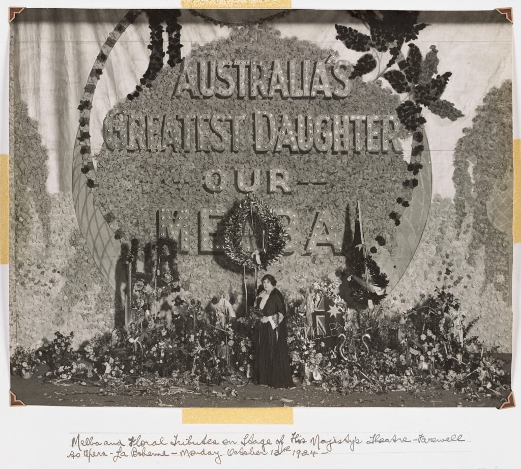 Melba standing in front of floral tributes with the text 'Australia's greatest daughter our Melba'