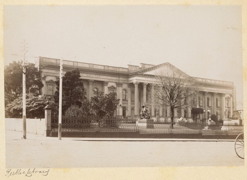 Sepia image of State Library Victoria in 1895, showing wrought iron fence and trees in forecourt.