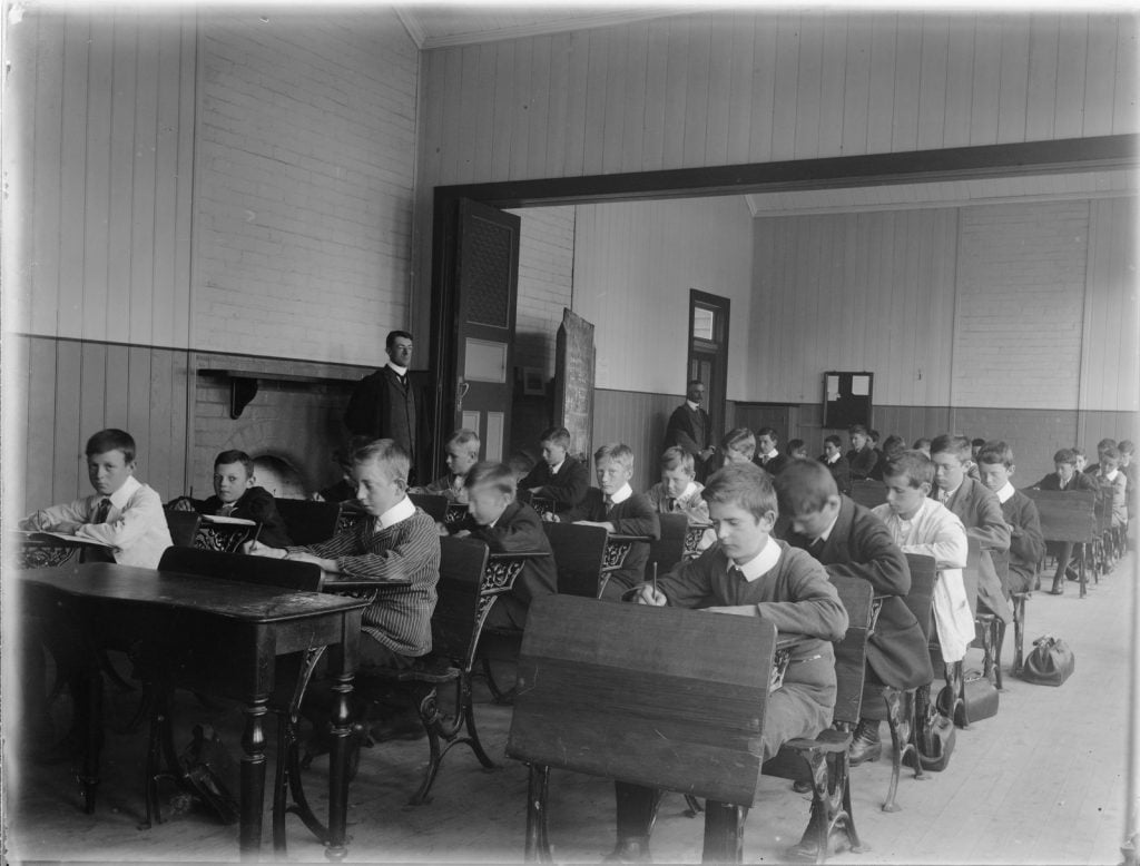Interior of classroom, with boys sitting at wooden desks.