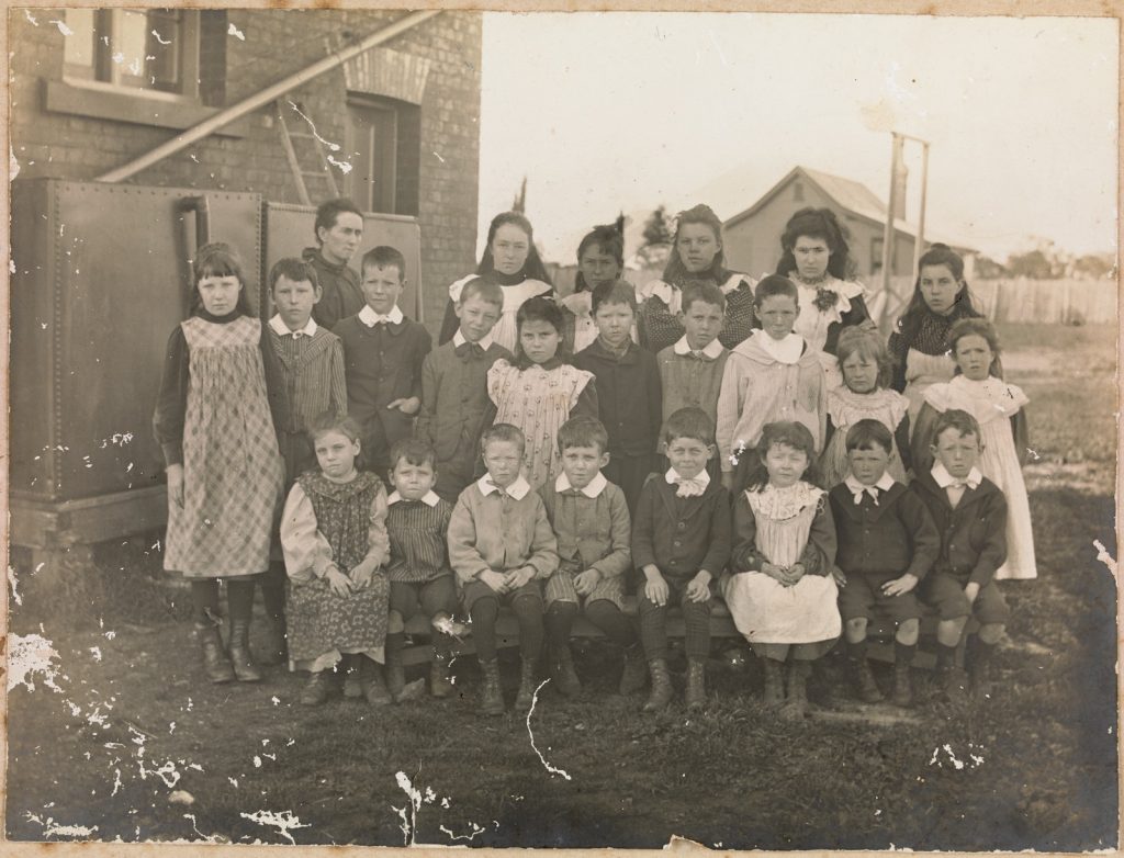 Group portrait of school children, front row seated, back two rows standing, teacher standing back left, with a brick building on left and another building in the background. Written on verso: Teesdale Primary School. c. 1900. Martha Rutter - Teacher.
Written on verso: Back Row: Harriet Cations, Ruby Murrell, Unknown, Gerty Miller, Violet Murrell. Middle Row: Vera Squires, Leroy Murrell, Tom Griffin, Ernest Squires, Mary Miller, Clifford Griffin, Robert Cations, Lillian Grant, Daisy Griffin. Front Row: Susie Miller, Robert Bull, unknown, Arthur Cations, ? Miller, Mary Flanagan, John King, Tom King.