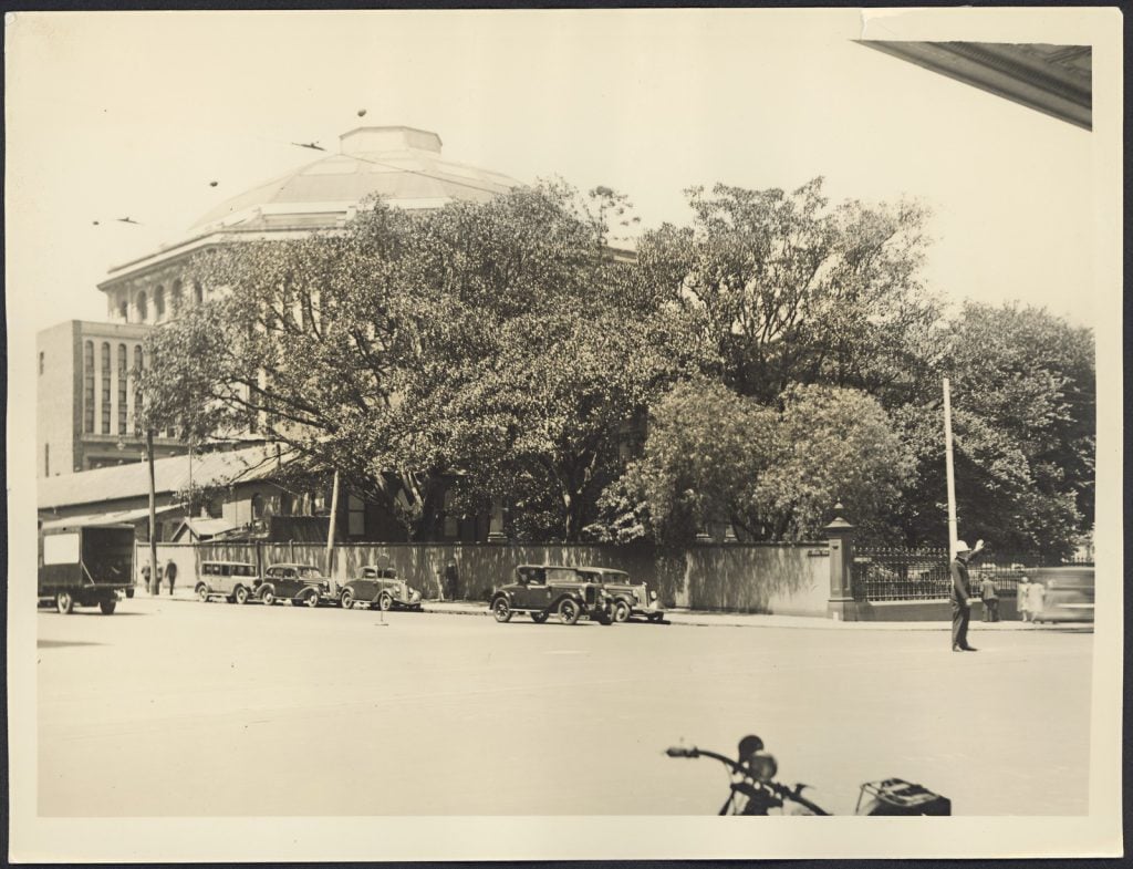 Sepia photograph of North-west corner State Library of Victoria prior to removal of fence & trees, 1938. Shows cars parked outside Library and a traffic policeman directing traffic.