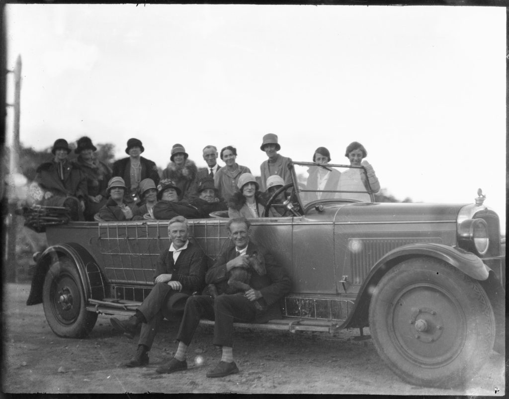 Black and white photo of a group of women in an open touring car, with two men, one with a dog, seated on the running board