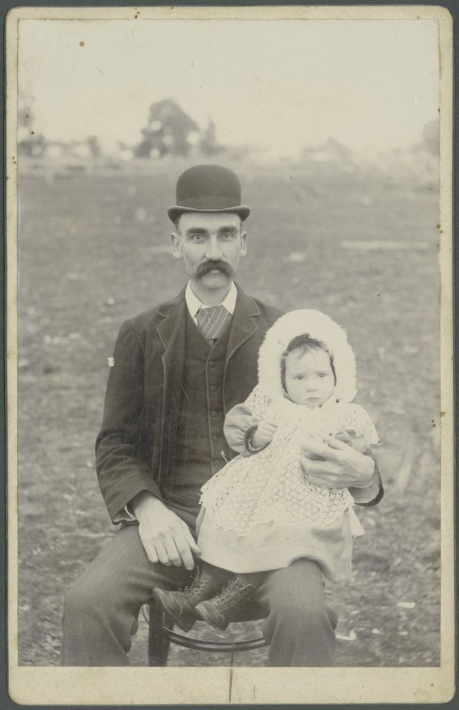 Black-and-white photograph of man with large moustache wearing bowler hat and suit, sitting on chair in a field, with young infant wearing white dress on his knee.