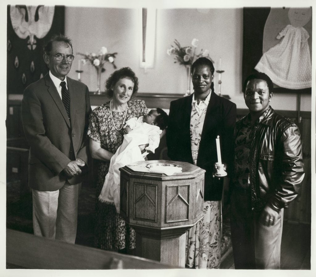 Black-and-white informal portrait of four people standing inside a church behind baptismal altar. Woman, centre, holds baby in baptism dress. Man, far right, holds lit candle.
