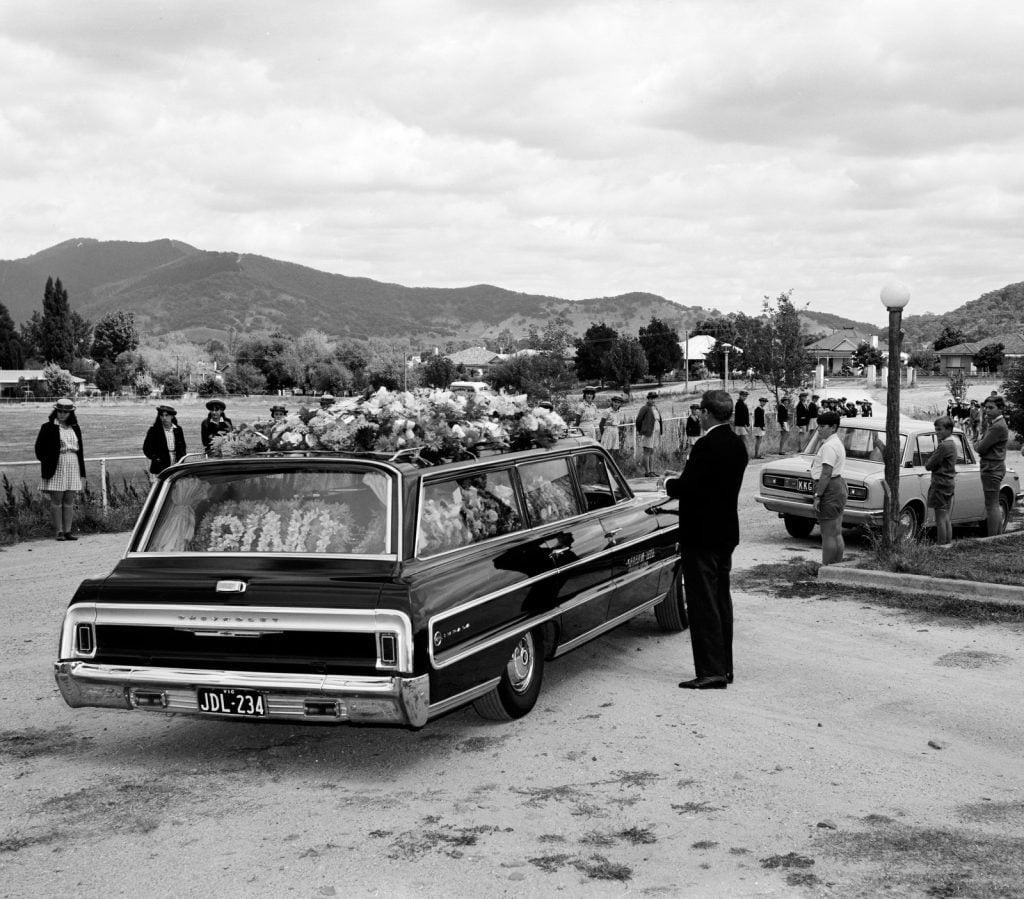 Black-and-white photograph of cars in wedding procession entering a cemetery, with onlookers standing either side. Hearse covererd with flowers closest to camera.