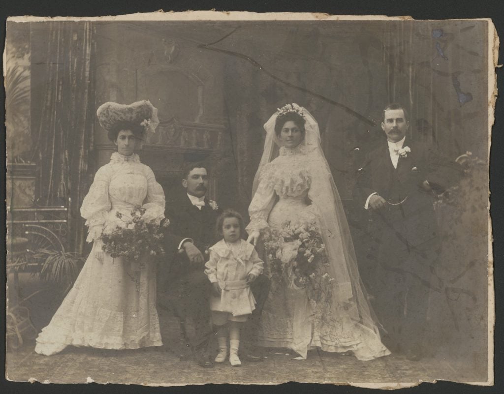 Black-and-white studio portrait of bride, groom, small child and two other people, most likely family members. Women both wear full-length white gowns, one in a lace hat; the bride in a veil.