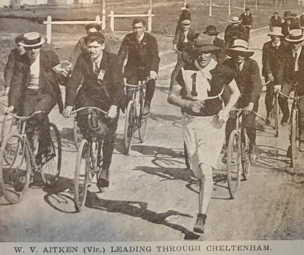 Photograph of a runner wearing a wide brimmed hat surrounded by several other men riding bicycles.