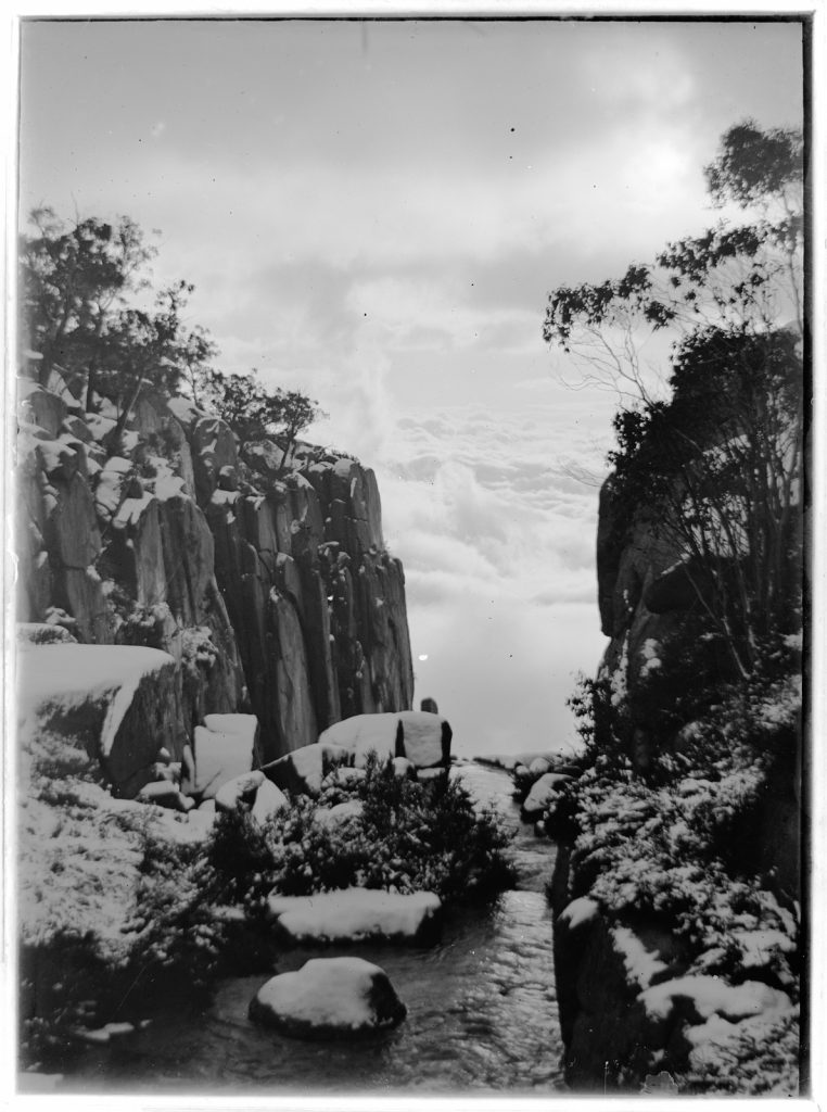 View through a snow covered ravine, with water running over the rocks, high looking over banks of clouds, with sun shining.