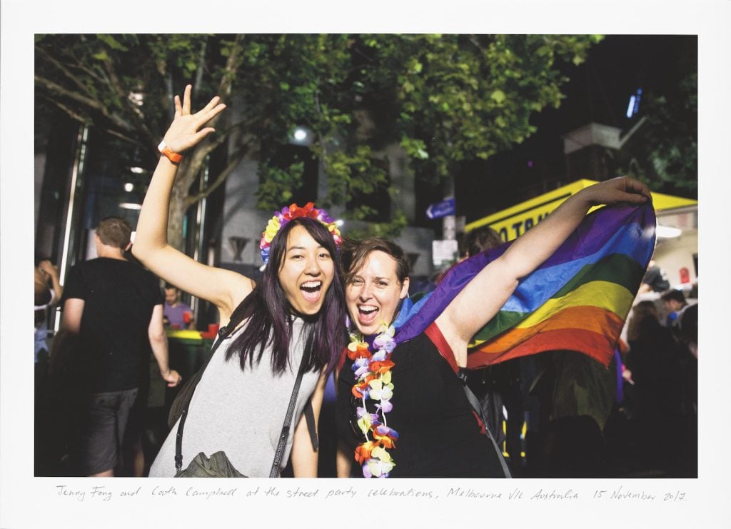 Two women in front of the State Library of Victoria at a street party celebration. Both women are smiling and wearing floral garlands. One woman is holding a rainbow flag.