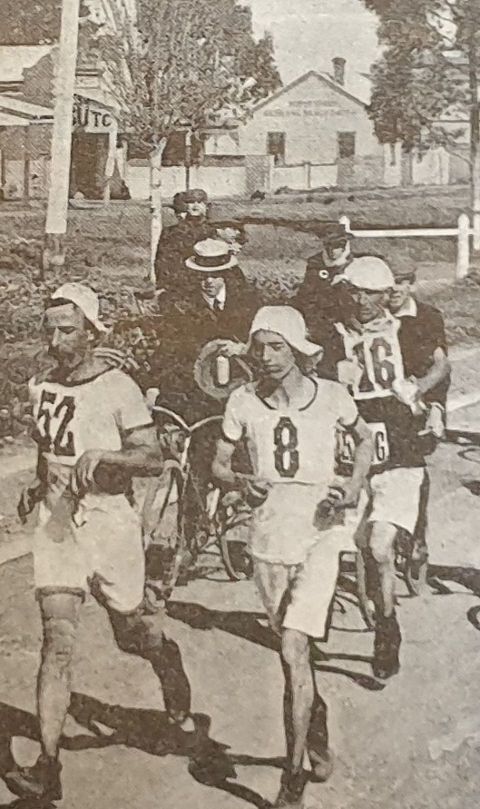 Photograph of three runners running on an unsealed road being followed by men on bicycles.