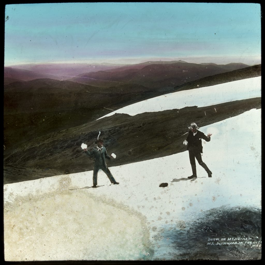 2 men throwing snowballs at each other, standing in a patch of snow,with bare receding hills, in the background. A sunny day, and the photo is colourised.