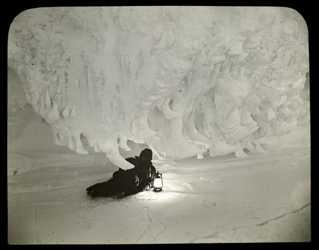 man dressed in snow proof gear holding a lantern, reclining on the snowy ground, with an icy ceiling