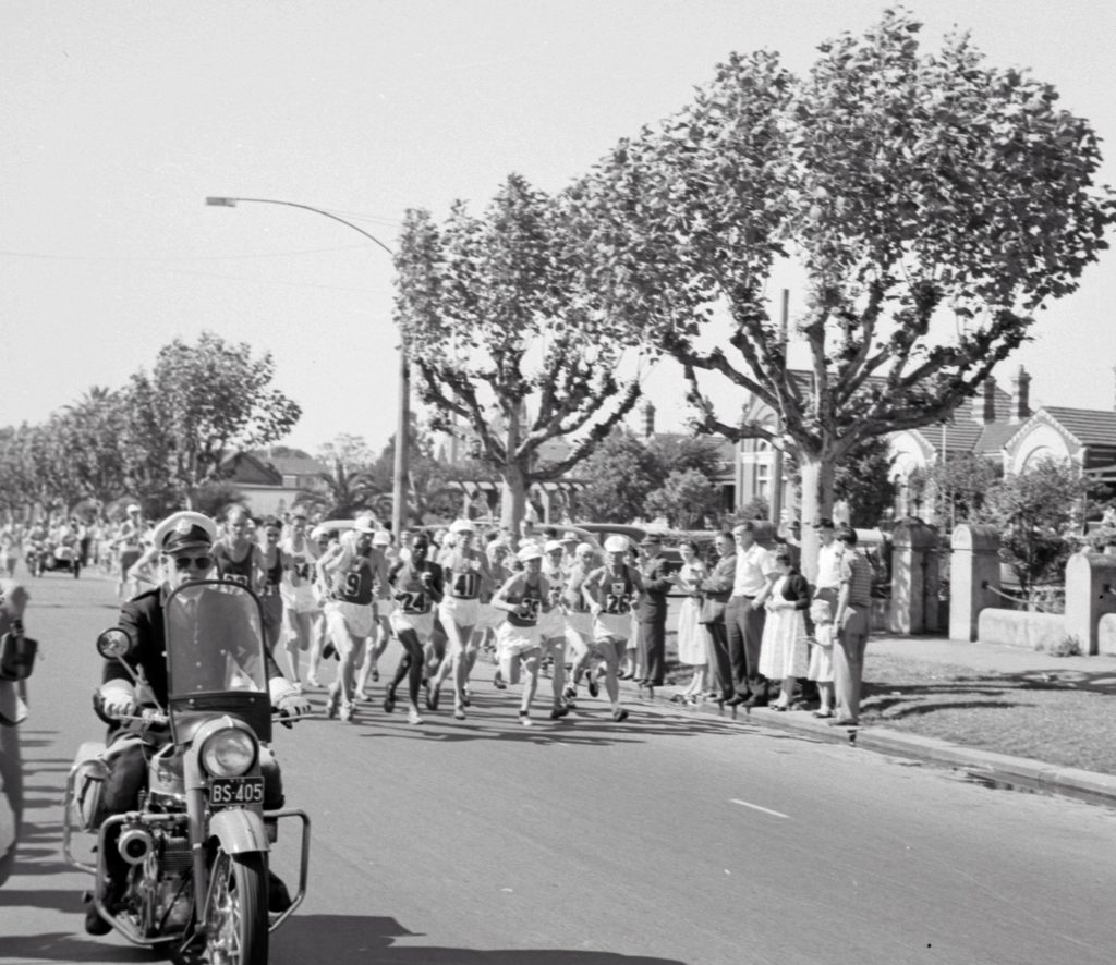 Group of runners on a road. Motorcycle rising in front of them and spectators standing on the nature strip. 