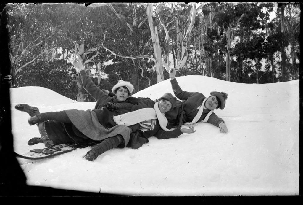3 young women tumbling about in the snow, falling of a sled , wearing hats, and scarves