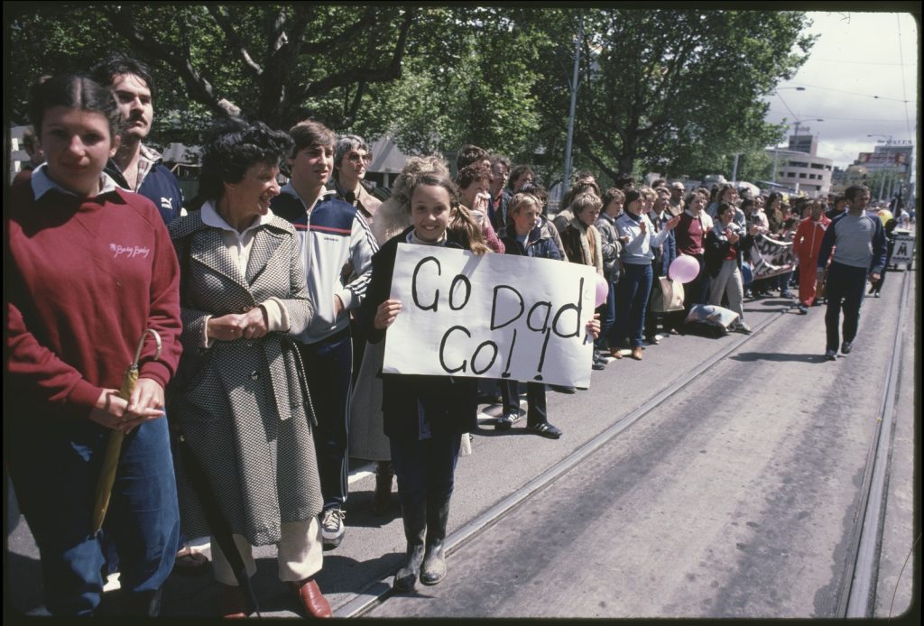 Photograph of a young woman spectating at a marathon, holding a handwritten sign saying 'go dad, go!!'