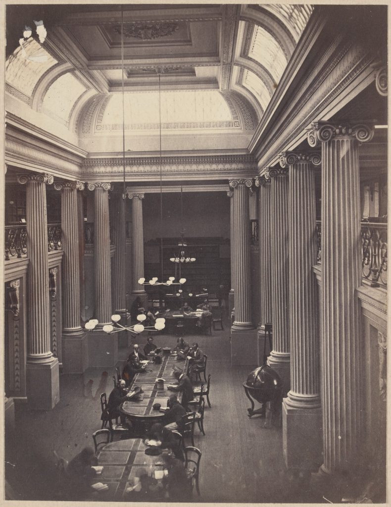 Queens Hall, the State Library reading room, the ceiling supported by  pillars, the room lit through sky lights, tables down the centre of the room, men reading, lights suspended from the double story ceiling, and a globe in a stand.
