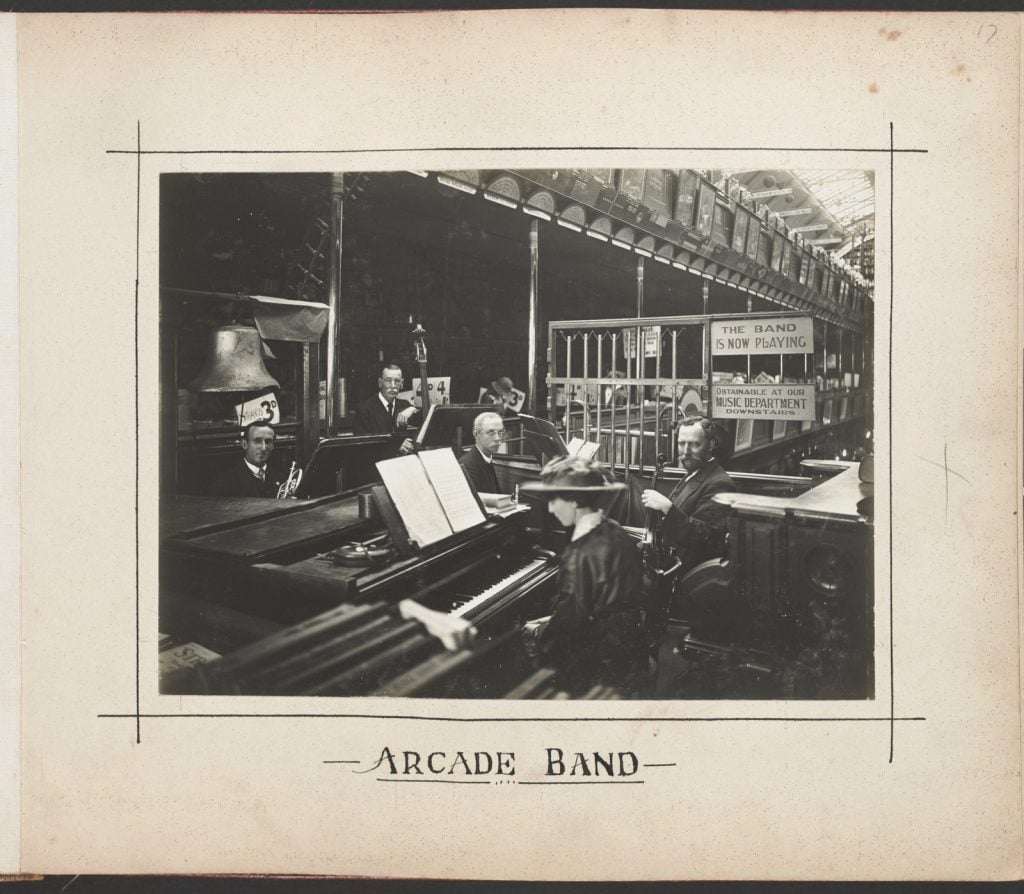 Black and white photograph showing band set up in the arcade in front of sign reading ‘the band is now playing’.
