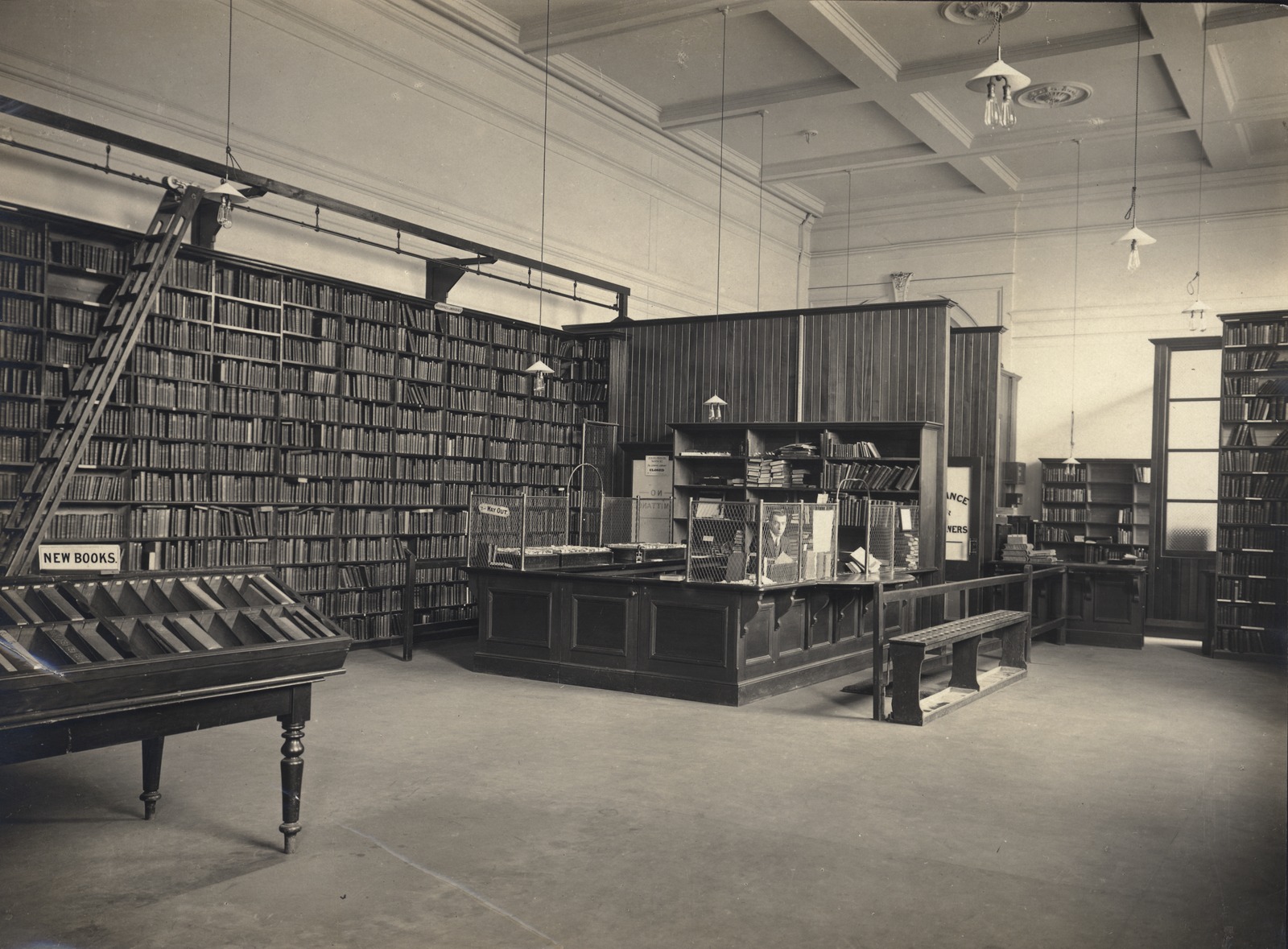 interior of the lending library at the state library, a wall of book shelves with a ladder on rails to access upper levels, a desk with a man behind it.