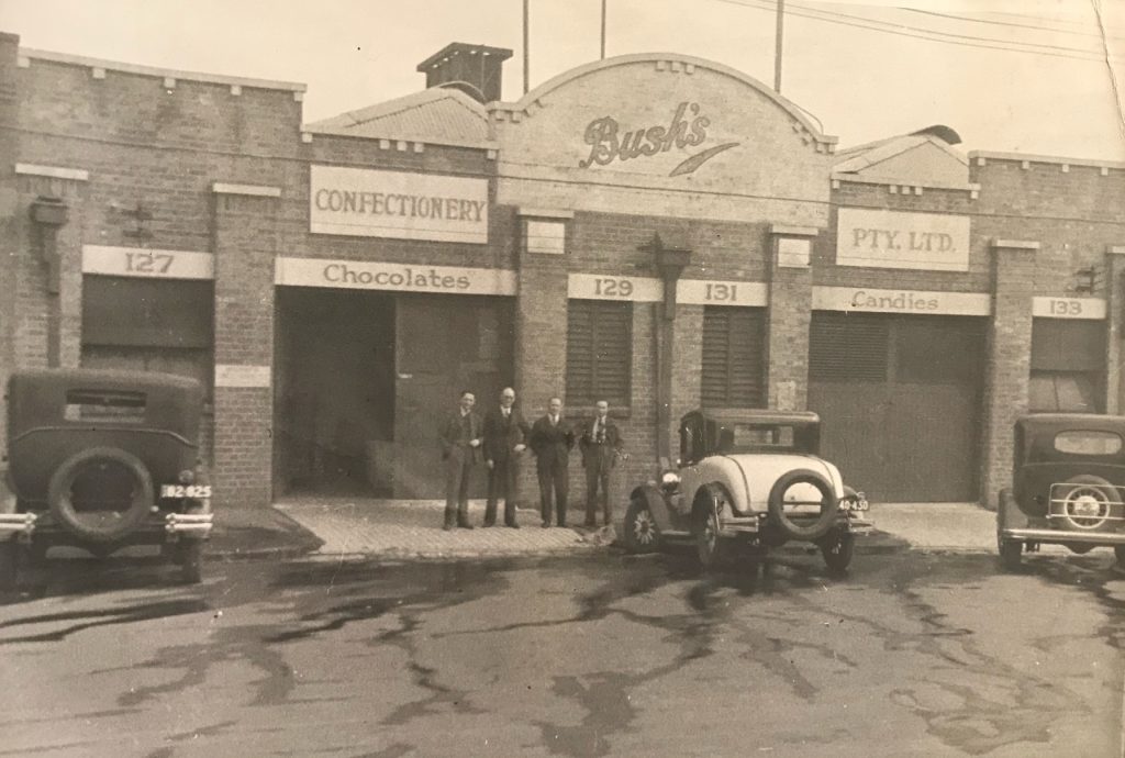 Bush's confectionery company factory, with four men standing at the front of the building, and three cars parked outside.