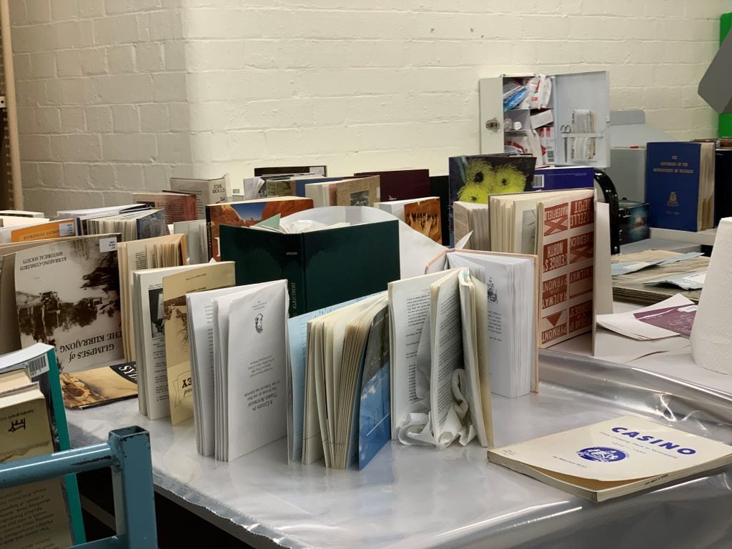Water-damaged books placed vertically on their spines on a tabletop. The books are fanned out to facilitate drying of the pages. Some have sheets of paper towel tucked between pages.