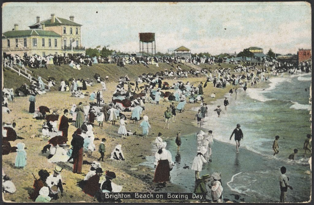 Well dressed people on the beach, with some in the shallow water. Large three story building in the background. 