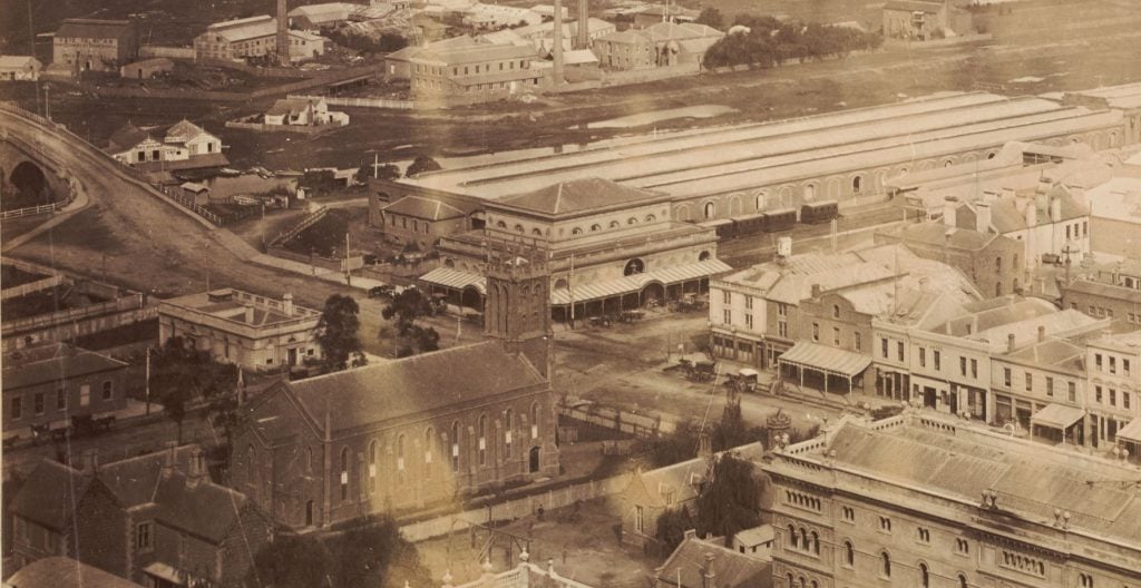 Elevated view showing St Paul's Anglican Cathedral on the intersection of Swanston and Flinders Streets, the fish market later replaced by Flinders Street Railway Station, railway bridge across Yarra River, other buildings, some identified: McNaughton & Co. (lower left), Princes Bridge Hotel (Flinders/Swanston Streets), McEwan & Compy., A.J. Smith C.F. Creswell, (Swanston Street); Emerald Hill (later South Melbourne) and bay in distance.