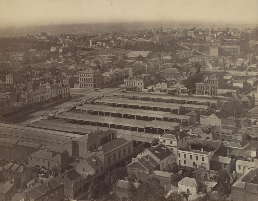 Photographic print, elevated view, of Melbourne looking east. Shows empty market shelters, hotels and houses, with other buidings including several churches in background.