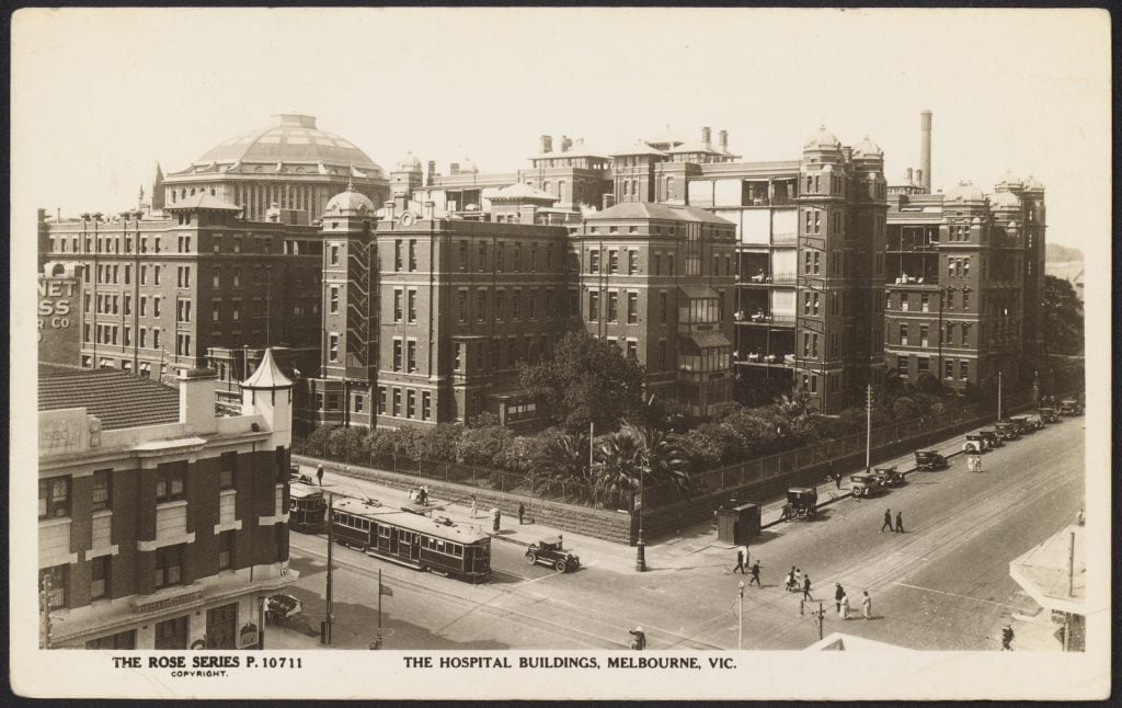 Elevated view, from corner of Lonsdale and Swanston Streets, of the old Queen Victoria Hospital, with dome of the State Library in left background. Cars at curb, horse and buggy, trams in Swanston Street.