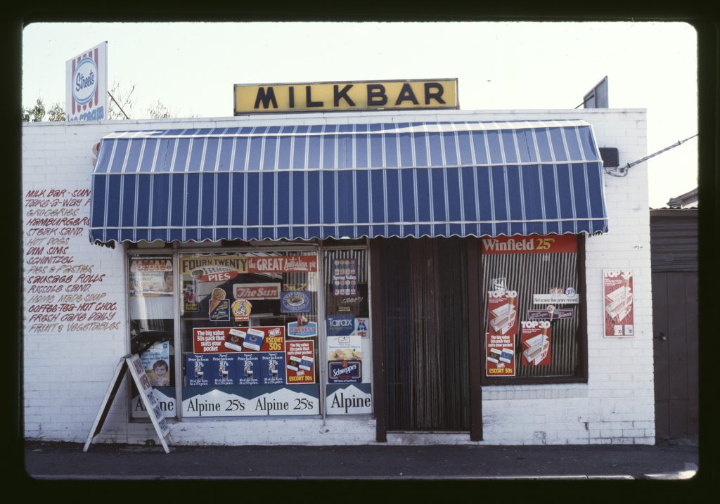 Store front with striped awning over front door and display window, Milk Bar sign at roof, advertising covering most of window