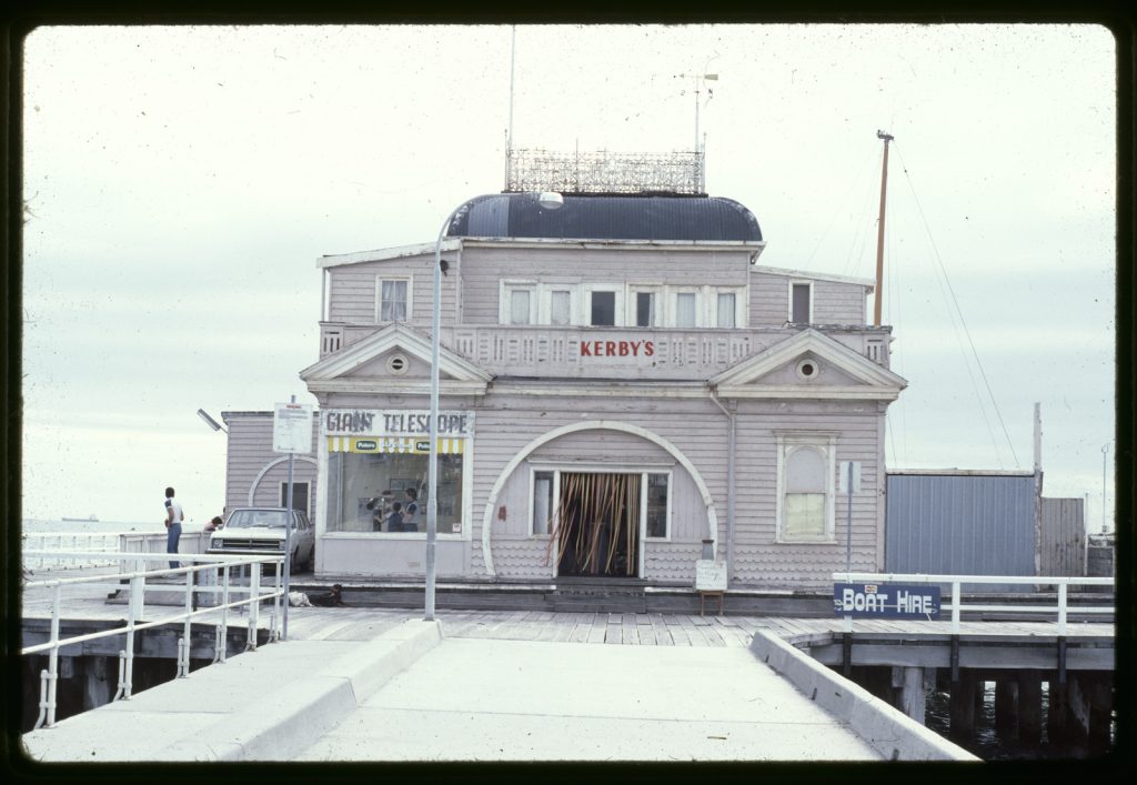 Kiosk at the end of the St. Kilda pier, the name Kirby's on wall below roof, plastic strip curtain across door.