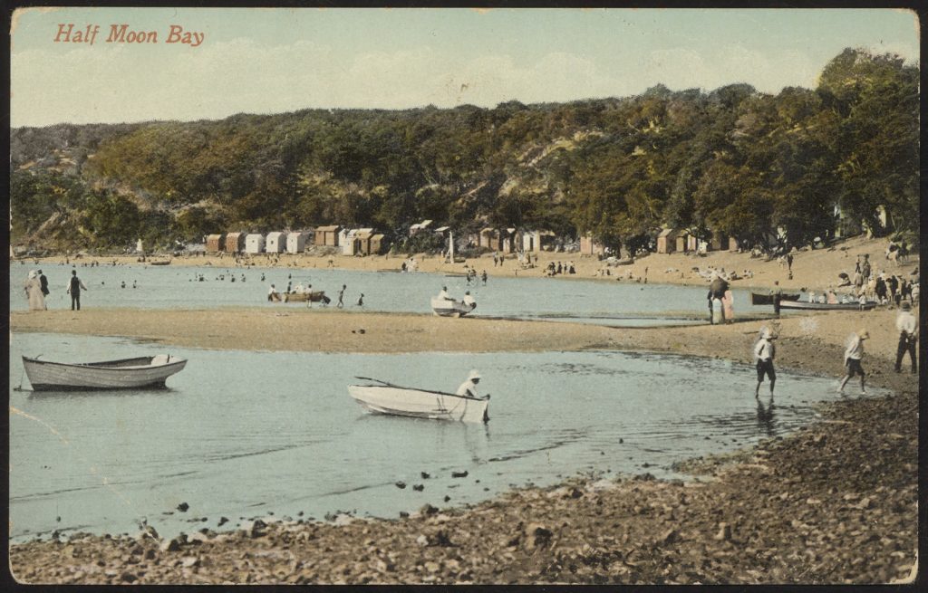 People at the beach. Sandbar extending into the water, some small boats, and wooden bathing boxes in the background. 