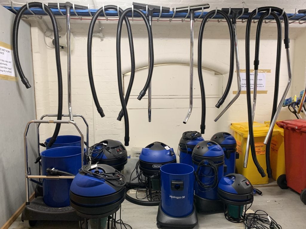 Around eight blue and black wet and dry vacuums in storeroom. Hoses are detached and hanging above vacuums which are sitting on the floor.