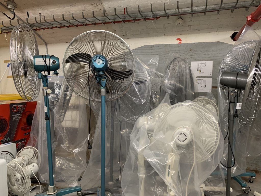 Large metal and plastic floor fans in storeroom. Some are covered with clear plastic, others are not.