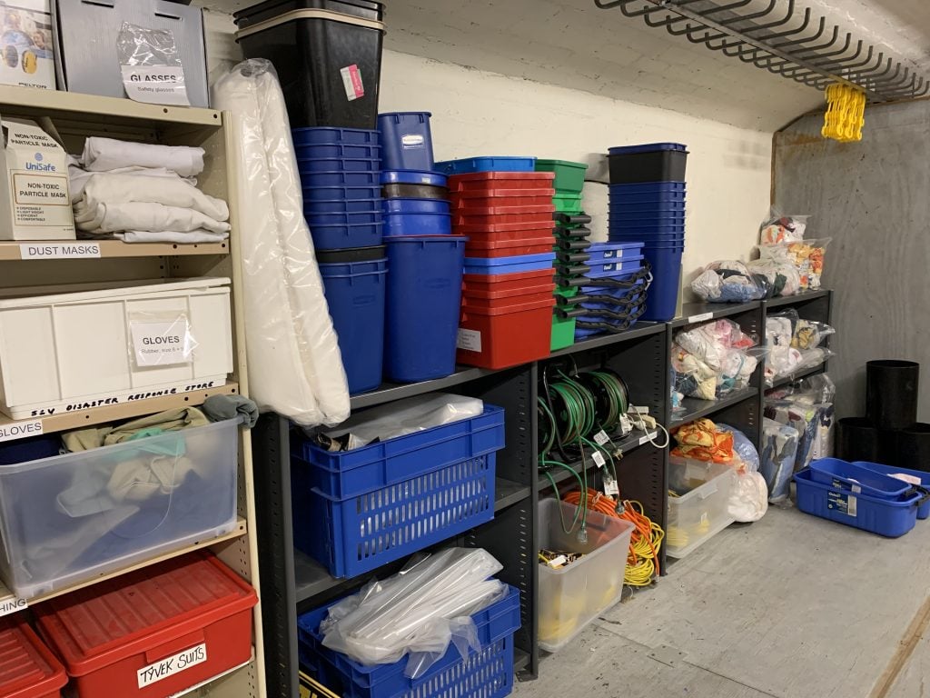 Shelving in a storeroom with disaster response supplies including various coloured plastic buckets, electrical extension cords, bags of rags, gloves and dust masks.