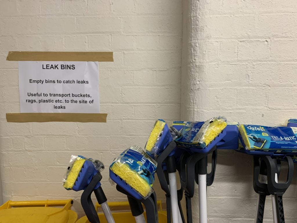 Sign taped to a cream-coloured brick wall which reads, 'LEAK BINS, Empty bins to catch leaks, Useful to transport buckets, rags, plastic, etc. to the site of leaks.' Also visible are the heads of six brand new, still in packaging, upturned Oates brand floor mops. These are blue and yellow.