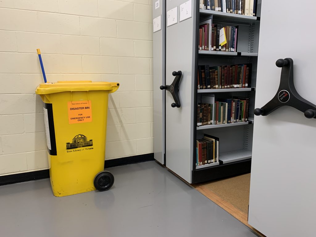 Large yellow wheelie bin, labelled 'Disaster Bin, State Library Victoria' located in collection store. Grey metal compactus shelving to the right of the bin contains books.
