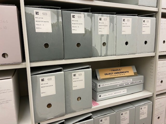 Blue grey cardboard archive boxes on grey metal shelving. The boxes are labelled, identifying them as collection material of State Library Victoria. Some of the boxes are partially wet due to a leak.