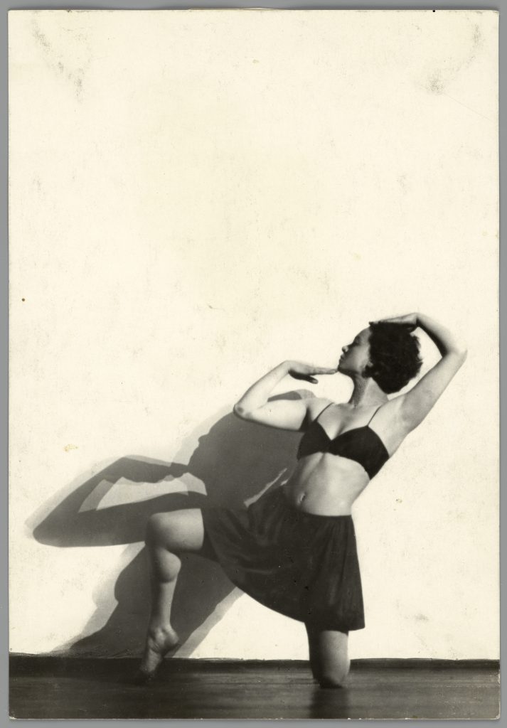 Black and white studio portrait of Sonia Revid, posing on one knee with her head turned into profile and her arms framing her face. Sonia wears a cropped bra-style top and skirt.