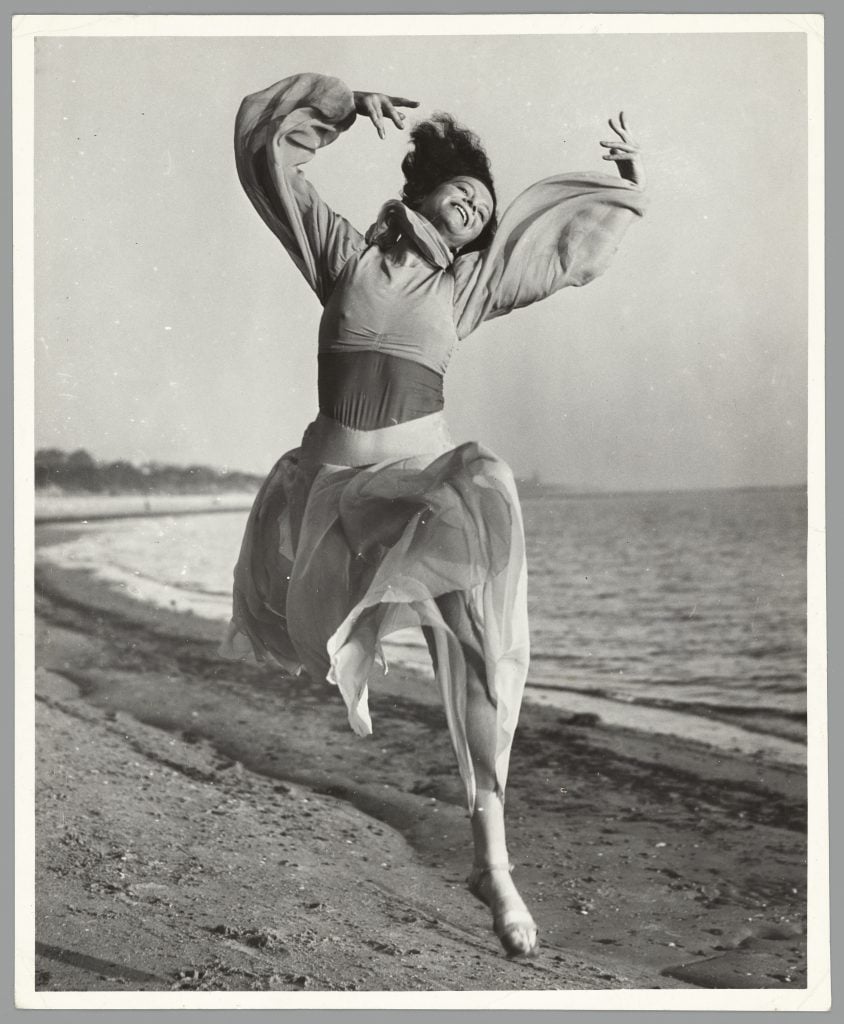 Black and white portrait of Sonia Revid, smiling, wearing a flowing white tule dress captured mid-leap on St Kilda beach.