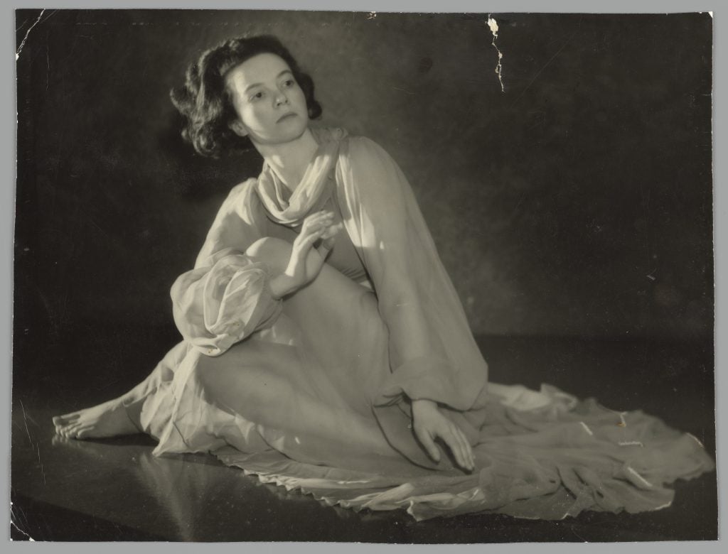 Black and white studio portrait of Sonia Revid in a long white dress made of tule. Sonia sits on the floor in a dancer's pose with her skirts arranged next to her.