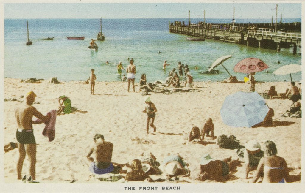 Colour view of Portsea beach with sunbathers on the sand, some under umbrellas. 