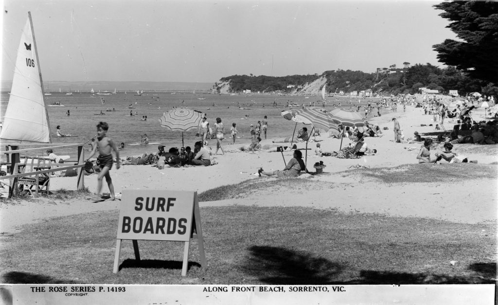 Beach with sunbathers and umbrellas. Sign in the foreground saying 'surf boards'.