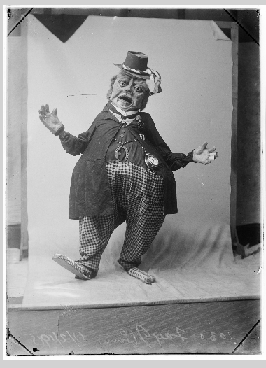 Unknown figure in fancy dress. 
Whole-length, standing, entire boy encased in fat clown costume with checked pants and large checked shoes, long coat buttoned at top button, horse shoe across waistcoat, alarm clock in place of a fob watch; also wearing a large papier mache head with a hat.