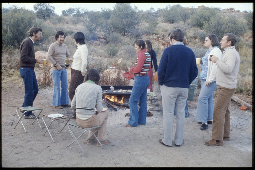 A group of young people standing around a bush barbeque with meat being cooked.