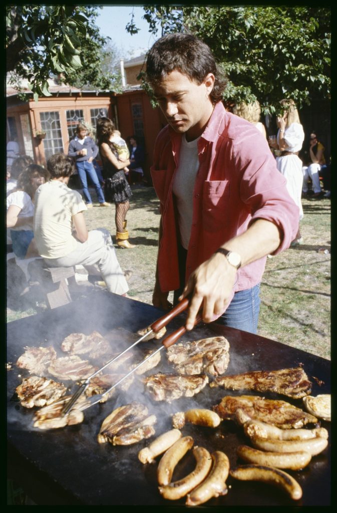 Shows a young man standing at a barbeque with sausages and chops, people in a backyard behind him.