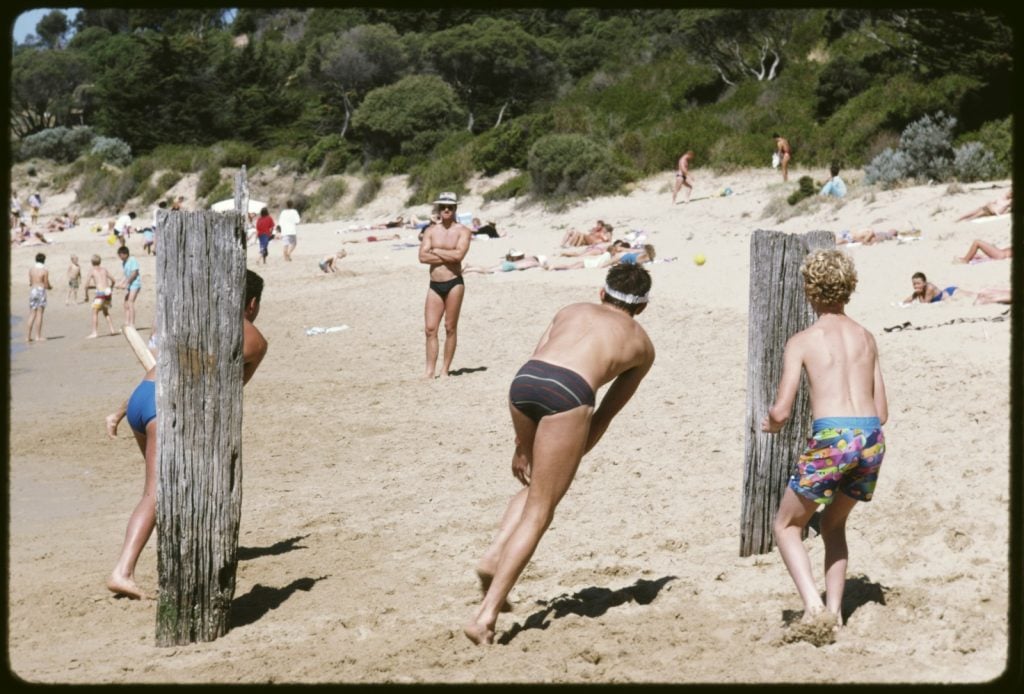 Men and boys, wearing bathers, playing beach cricket on the sand at Portsea Beach. They are using old pier piles as wickets. On the beach in the background, beachgoers are sunbathing and relaxing. Coastal vegetation, inclugind tea-tree, forms a backdrop to the picture.