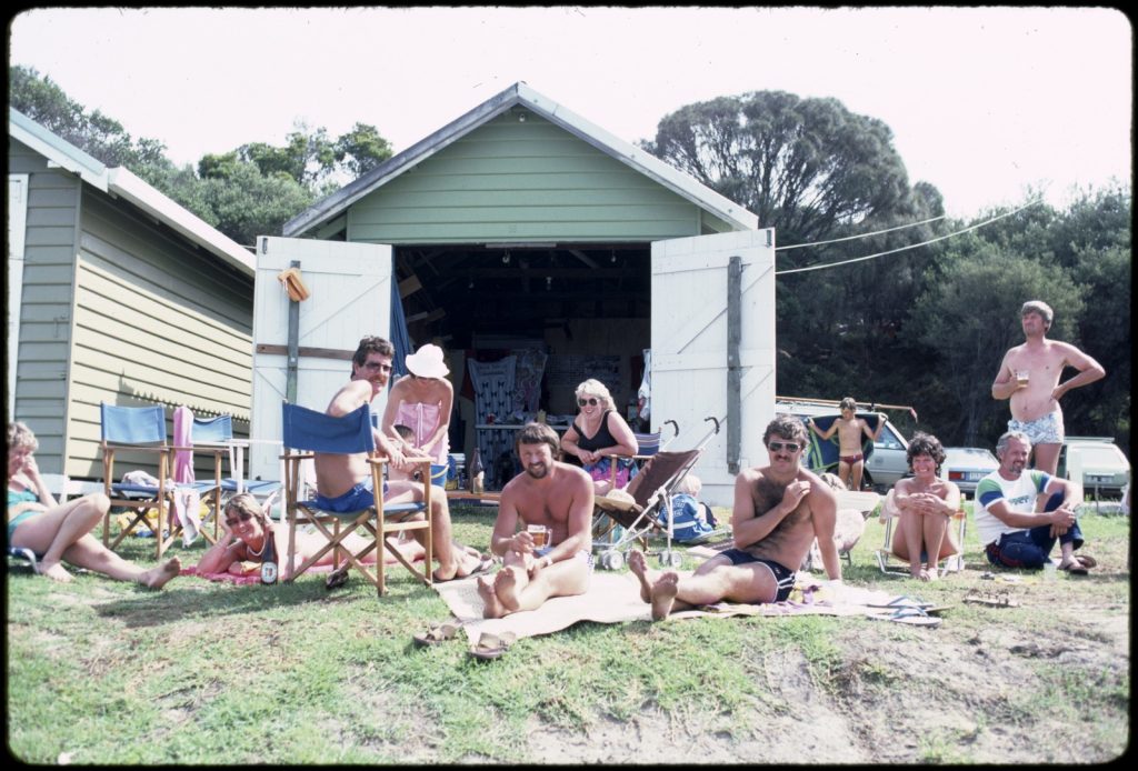Men and women in bathing suits, some seated on beach towels or deck chairs, beach hut with doors wide open in background.